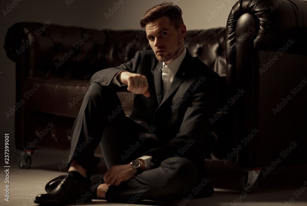 Businessman in a suit and a shirt is sitting and pondering next to the leather brown sofa.