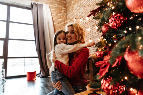 Christmas. Childhood. Home. Little girl and her mom are standing near the Xmas tree, hugging, looking at camera and smiling