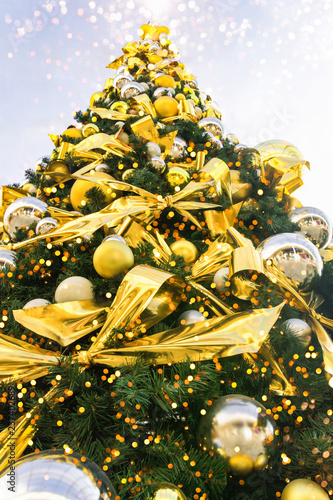 Big beautiful Christmas tree decorated with ornaments golden silver hanging balls bows garland sparkling lights in european city. Festive magic atmosphere. Low angle perspective. Poster greeting card