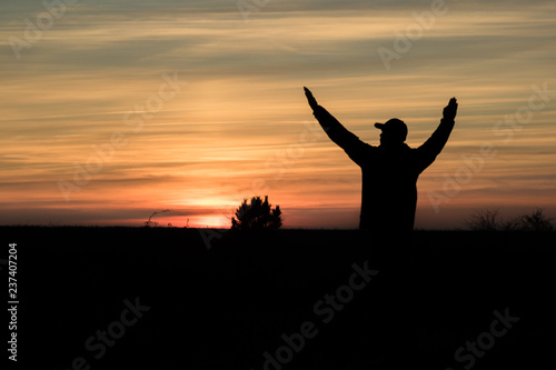 man admiring the red autumn sunset in the field and holding hands raised, the silhouette of a man and a tree
