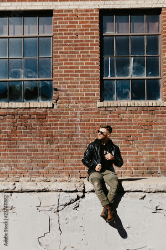 Young Attractive Modern Fashion Caucasian Male Guy Walking, Sitting, Smiling, and Laughing Outside Urban Old Abandoned Brick Warehouse Building in Urban City in Winter Season on Bright Sunny Day