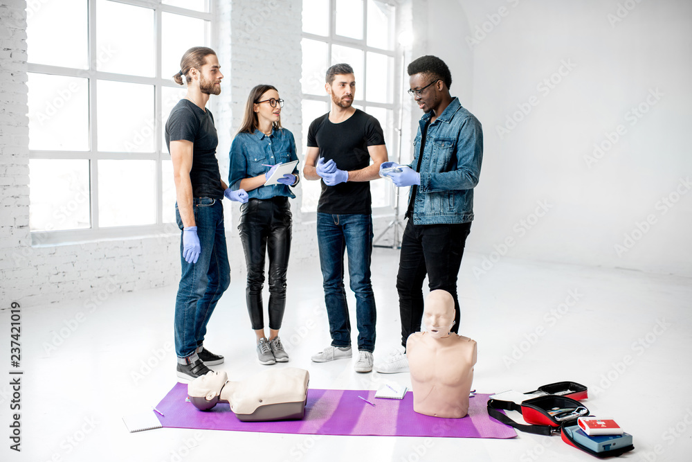 Fototapeta premium Young people standing and talking together during the first aid training with medical stuff and dummies on the floor