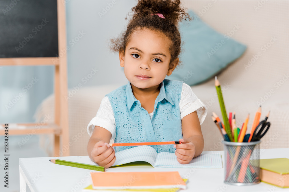 adorable african american kid holding pencil and looking at camera while writing in workbook adorable african american kid holding pencil and looking at camera while writing in workbook
