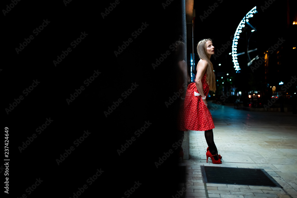 Fototapeta premium A beautiful mature woman in a red dress standing on the street and leaning to the wall at night with the city lights in the background.