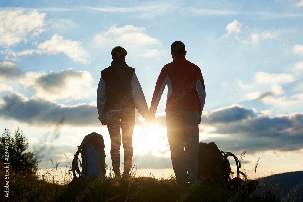 Back view of young persons admiring the sunset in the mountains holding hands. Near them there are backpacks. The sun is visible between their silhouettes