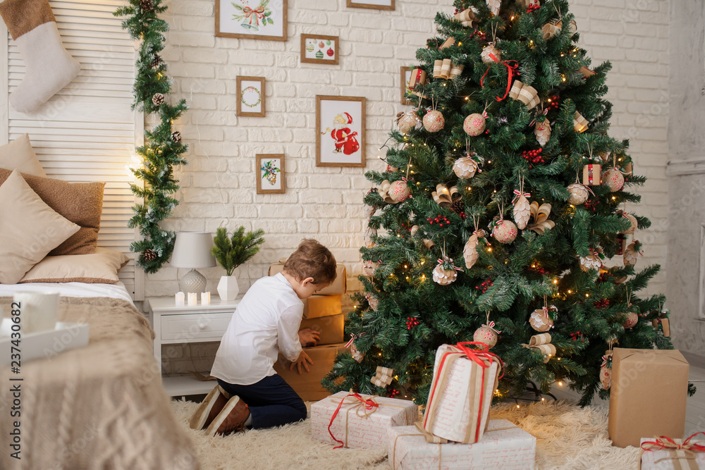 a 6-year blond boy with a present in his hand near a christmas tree and a bed a 6-year blond boy with a present in his hand near a christmas tree and a bed