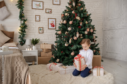 a 6-year blond boy with a present in his hand near a christmas tree and a bed