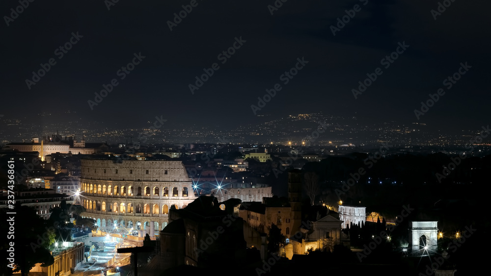Panoramic night view of Rome. In the foreground the illuminated Colosseum, on the right the Arch of Constantine and the area of the Imperial forums with the Arch of Titus