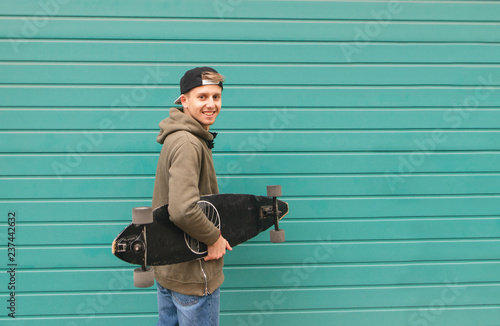 Happy student with a longboard in his hands on a color turquoise background, looking into the camera and smiling. Portrait of a positive skateboarder.