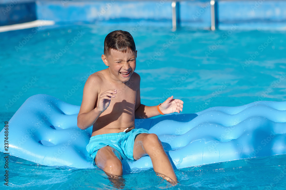  Caucasian boy is  sitting  on blue  inflatable mattress at hotel swimming pool. He is enjoying his summer vacations and smiling.