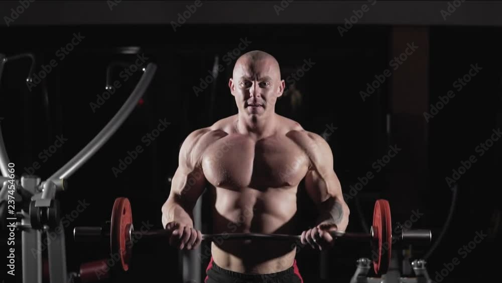 Bodybuilder doing exercise with barbell in dark gym. Exercises for muscles of biceps in slow motion. Weightlifter lifts barbell to training biceps. Medium shot Bodybuilder doing exercise with barbell in dark gym. Exercises for muscles of biceps in slow motion. Weightlifter lifts barbell to training biceps. Medium shot