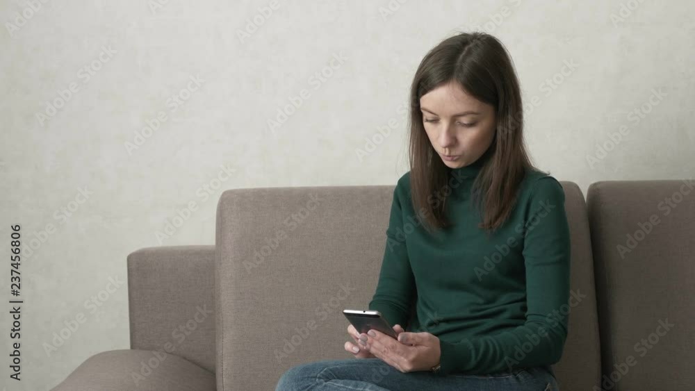 Attractive woman in green sweater and blue jeans using smartphone while lying on the couch in the living room. Typing, watching video, playing games on smartphone. Internet addiction concept. Attractive woman in green sweater and blue jeans using smartphone while lying on the couch in the living room. Typing, watching video, playing games on smartphone. Internet addiction concept.