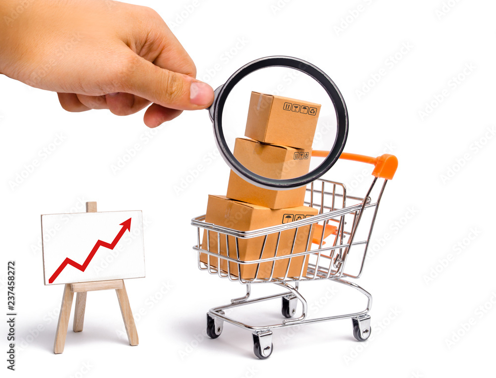 Magnifying glass is looking at the Supermarket cart with boxes and a graph with red arrow, merchandise: the concept of buying and selling goods and services, trade and turnover. Import and export