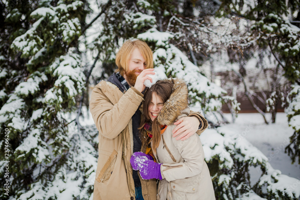 Young Caucasian boy with a beard and a girl have a date outdoors in the winter park against the background of a snowy conifer tree play snowballs, throw snow, hang themselves and play winter games Young Caucasian boy with a beard and a girl have a date outdoors in the winter park against the background of a snowy conifer tree play snowballs, throw snow, hang themselves and play winter games