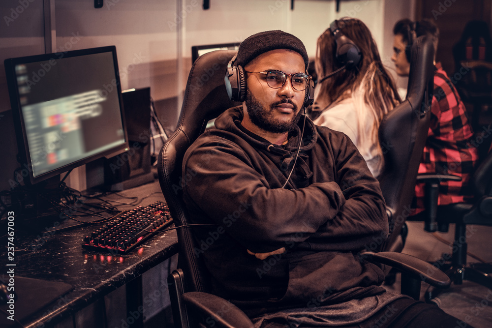 African-American bearded guy wearing hoodie and cap with her arms crossed sitting on a gamer chair in a gaming club or internet cafe.
