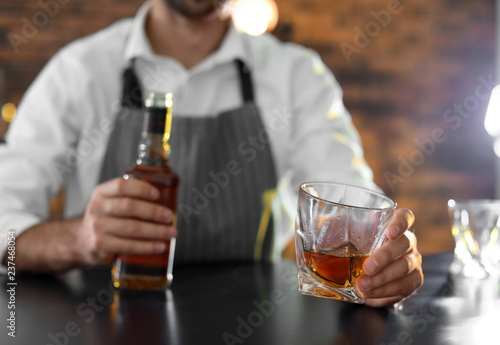 Bartender with glass and bottle of whiskey at counter in bar, closeup. Space for text