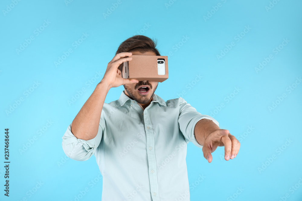 Fototapeta premium Young man using cardboard virtual reality headset on color background