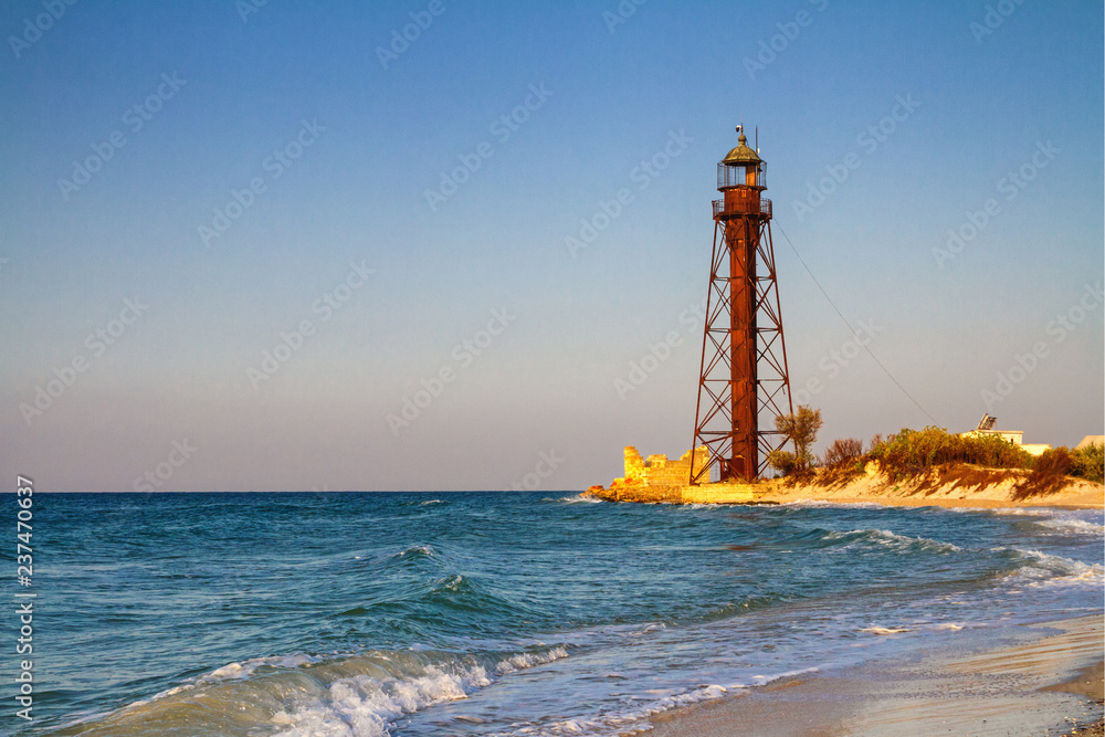 Old rusty abandoned lighthouse on the sea coast during sunrise near the ruins on sandy beach