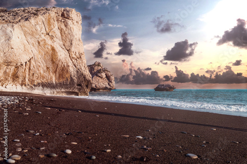 View at the Aprodite´s rock in Paphos, Cyprus with a cloudy sunset and the ocean with waves in the background.  Dark sand with pebbles and rocks is to be seen in the foreground.  photo