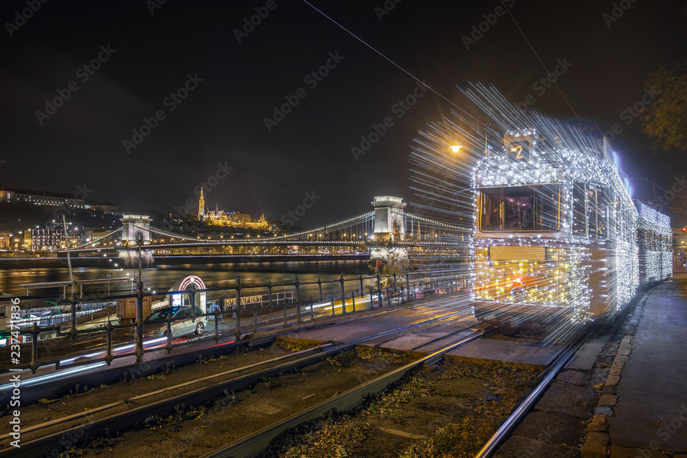 Obraz premium Budapest, Hungary - Festively decorated light tram (fenyvillamos) on the move with Szechenyi Chain Bridge, Matthias Church and Fisherman's Bastion by night. Christmas season in Budapest
