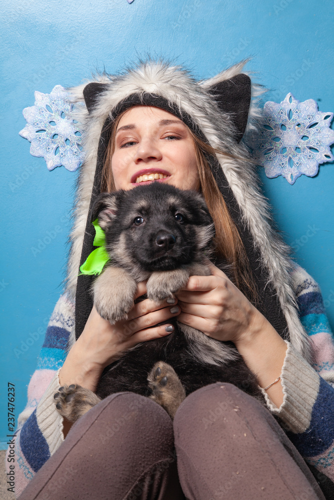 Obraz premium Pretty Woman In Fur Winter Hat Holding Little Cute shepherd puppy and smiling on blue background
