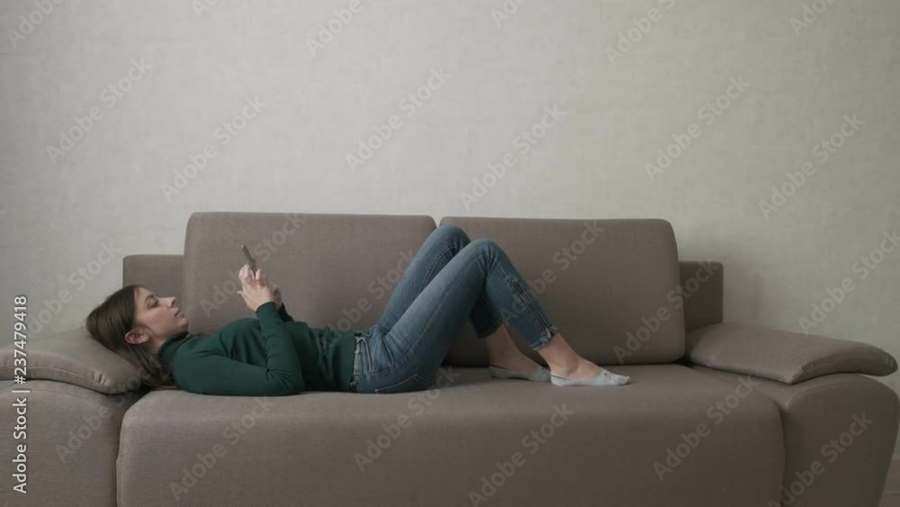 Attractive woman using smartphone while lying on the couch in the living room. Typing, watching video, playing games on smartphone. Internet addiction concept. Full shot. Attractive woman using smartphone while lying on the couch in the living room. Typing, watching video, playing games on smartphone. Internet addiction concept. Full shot.