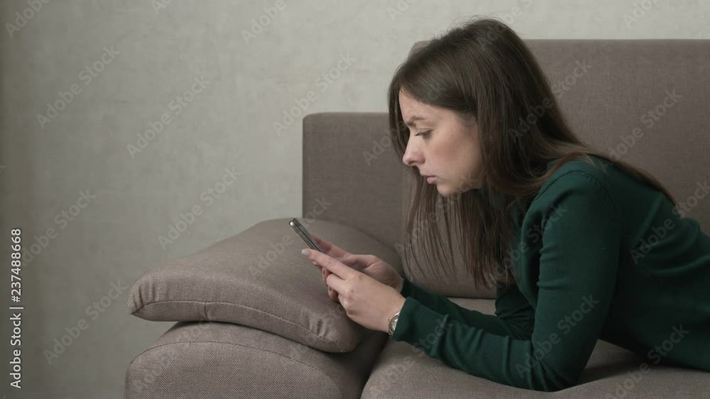 Attractive woman using smartphone while lying on the couch in the living room. Typing, watching video, playing games on smartphone. Internet addiction concept. Medium shot. Attractive woman using smartphone while lying on the couch in the living room. Typing, watching video, playing games on smartphone. Internet addiction concept. Medium shot.