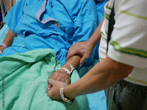 Elderly man s hands holding on a hand of an elderly female patient lying on a bed in a patient room at a hospital