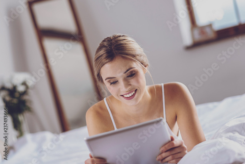Charming young woman lying on bed and texting on tablet