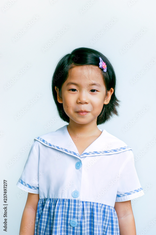 Happy little shy girl smiling at camera. Portrait of Thai pre-elementary school student in Thai uniform stand on white background and copy space. Learning, education or back to school concept. Happy little shy girl smiling at camera. Portrait of Thai pre-elementary school student in Thai uniform stand on white background and copy space. Learning, education or back to school concept.