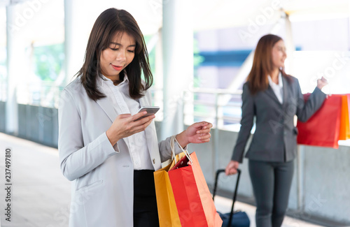Happy Asian girl holding bags with purchases, smiling while looking at phone in shopping center. Young business woman walking and reading message or using app on smartphone.