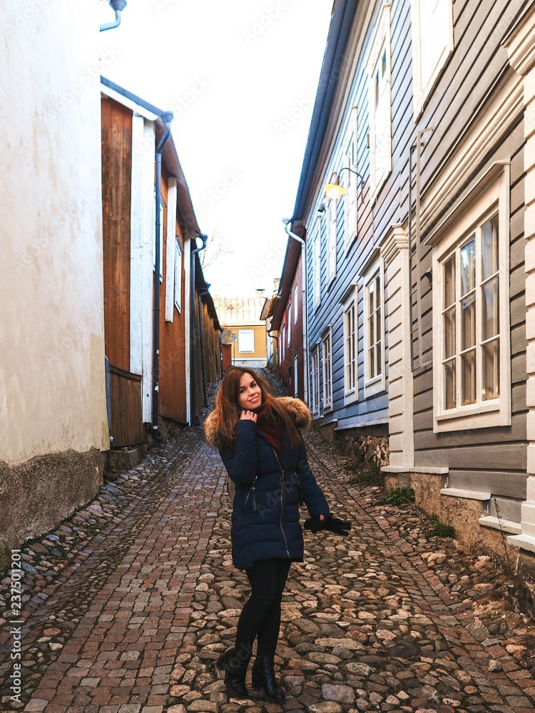 Brunette girl in winter jacket with scarf stands in the middle of a Finnish street with Scandinavian houses