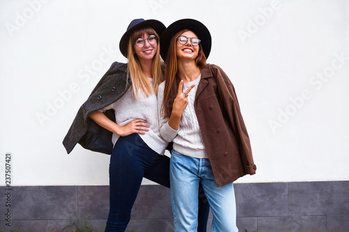 Positive lifestyle image of two exited hipster women posing over white wall,wearing jeans,hat, jacket and sweaters Fall season and spring. Smiling and spending great time together. Copy space.