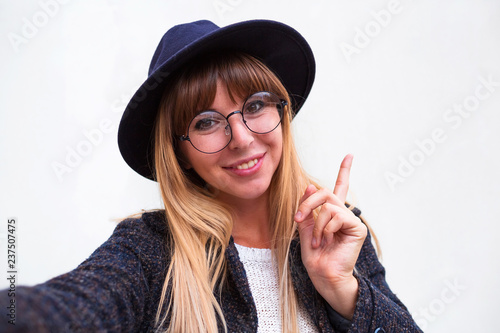 Happy woman in glasses with beaming smile making selfie and pointing finger up.Studio portrait of beautiful woman smiling with white teeth and making selfie, photographing herself over gray background