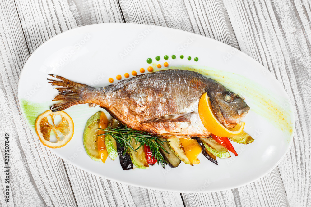 Grilled dorado fish with baked vegetables and rosemary on plate on wooden background close up. Healthy food. Top view Grilled dorado fish with baked vegetables and rosemary on plate on wooden background close up. Healthy food. Top view