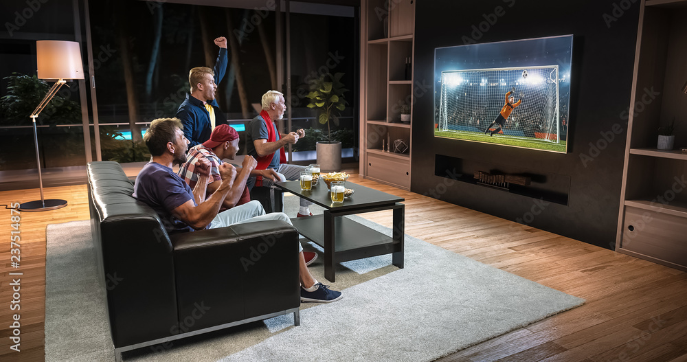 Group of fans are watching a soccer moment on the TV and celebrating a goal, sitting on the couch in the living room.