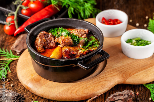 The concept of Serbian cuisine. Juicy baked beef in its own juice with potatoes, vegetables and greens. Serve in an iron fire on a wooden board.