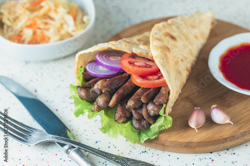 Handmade doner kebab is lying on the white table made of artificial stone Shawarma on the wooden cutting board with chicken meat  onions  salad lies on a dark old wooden table.