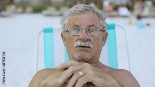 Close up portrait of senior man with moustache looking to the camera at the beach in deckchair 4k