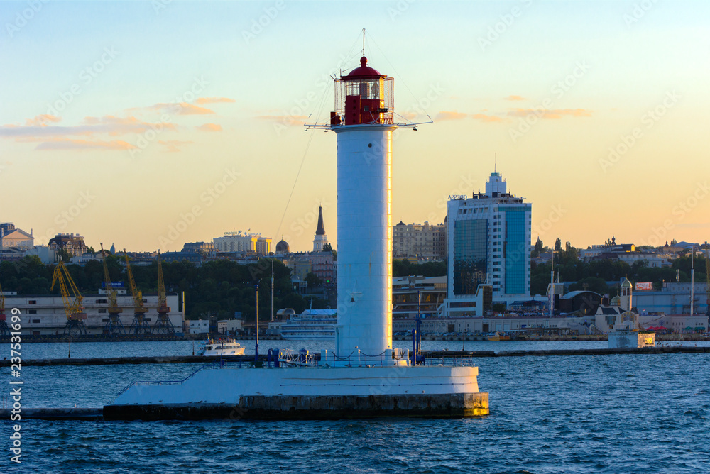 A lighthouse in the sea at the entrance to the port against the backdrop of a beautiful evening blue sky in soft colors. Summer seascape. White lighthouse with a red top. Black Sea. Seaport of Odessa A lighthouse in the sea at the entrance to the port against the backdrop of a beautiful evening blue sky in soft colors. Summer seascape. White lighthouse with a red top. Black Sea. Seaport of Odessa