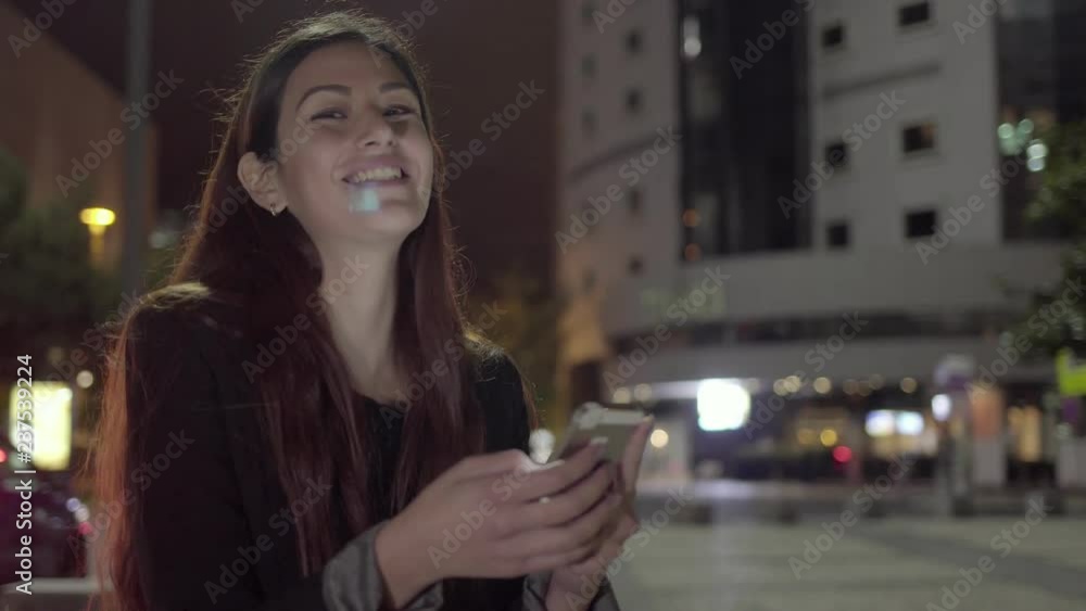 Cheerful young woman chatting on smartphone in evening city with cars. Beautiful happy girl networking and texting on smartphone outdoors. Using device concept Cheerful young woman chatting on smartphone in evening city with cars. Beautiful happy girl networking and texting on smartphone outdoors. Using device concept
