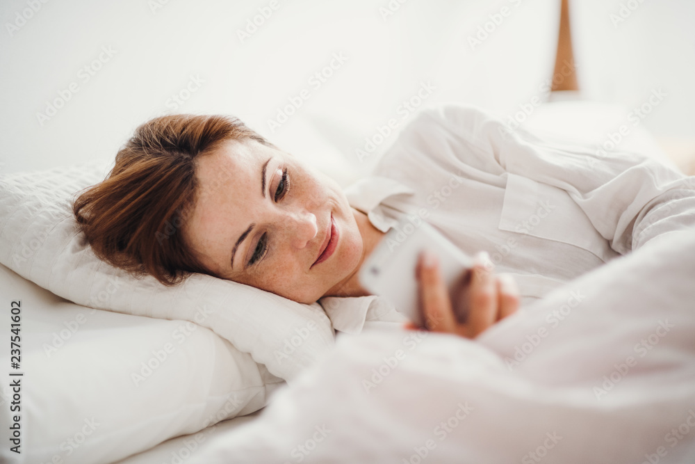 A young woman lying in bed indoors in the morning in a bedroom, using smartphone. A young woman lying in bed indoors in the morning in a bedroom, using smartphone.