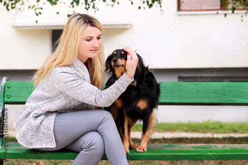 Beautiful woman with his small mixed breed dog sitting and posing in front of camera on wooden bench at city park. Portrait of owner and cute half breed dog enjoys, resting, petting together outdoors.