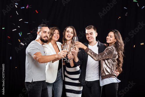 Company of friends in stylish casual clothes stand together and clink glasses with champagne on a black background and confiture around. Party time