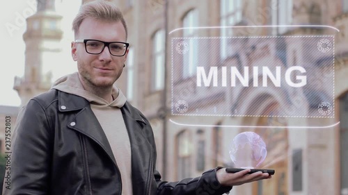 Smart young man with glasses shows a conceptual hologram Mining. Student in casual clothes with future technology mobile screen on university background photo