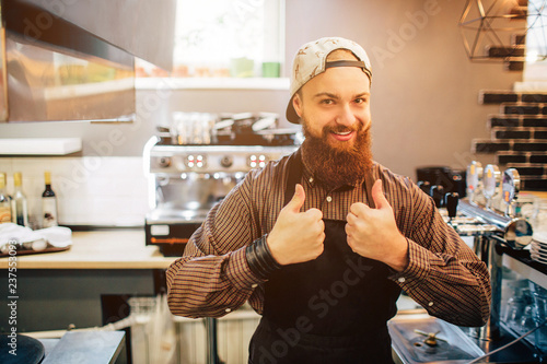 Happy young barista stand and show big thumb up on both hands. He look on camer. Guy stand in kitchen. Light comes out of window.