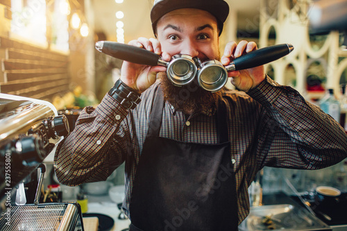 Another picture of happy and funny young barista holding to cezve and cover mouth with them. He looks on camera. Guy stand at coffee machine. photo