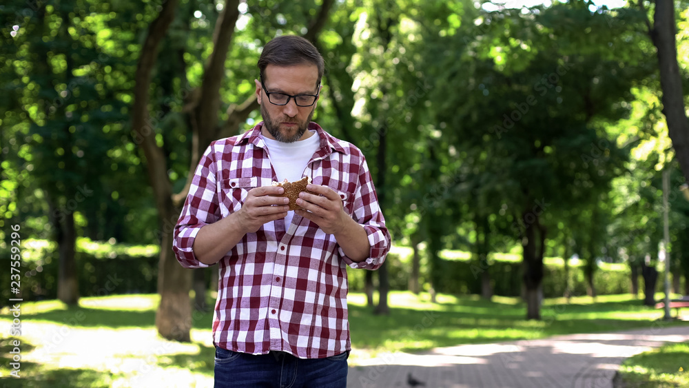 Guy chewing fast food burger in park feeling nausea, food poisoning symptom