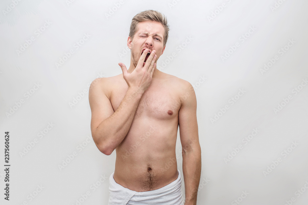 Sleepy young man stand and yawn. His hips is covered with white towel. Guy is shirtless. Isolated on white background.
