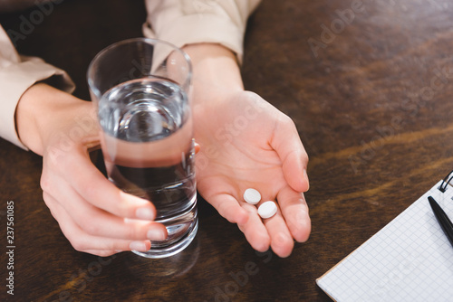 close-up partial view of businesswoman holding pills and glass of water at workplace